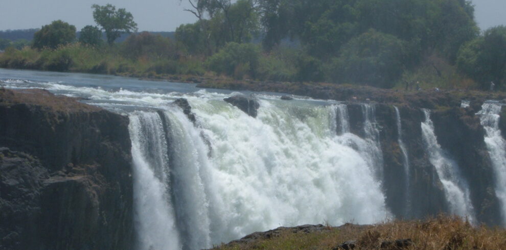 Vista del salto d'acqua delle Cascate Vittoria dal lato dello Zimbabwe, con la sponda opposta dello Zambia e bagnanti vicino al ciglio dello strapiombo.