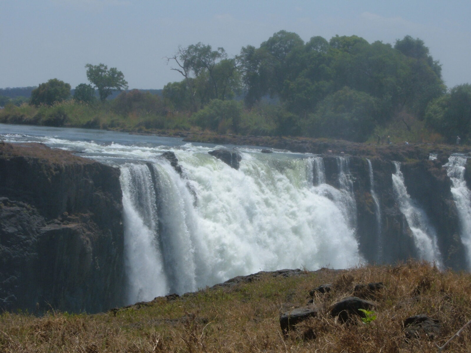 Vista del salto d'acqua delle Cascate Vittoria dal lato dello Zimbabwe, con la sponda opposta dello Zambia e bagnanti vicino al ciglio dello strapiombo.
