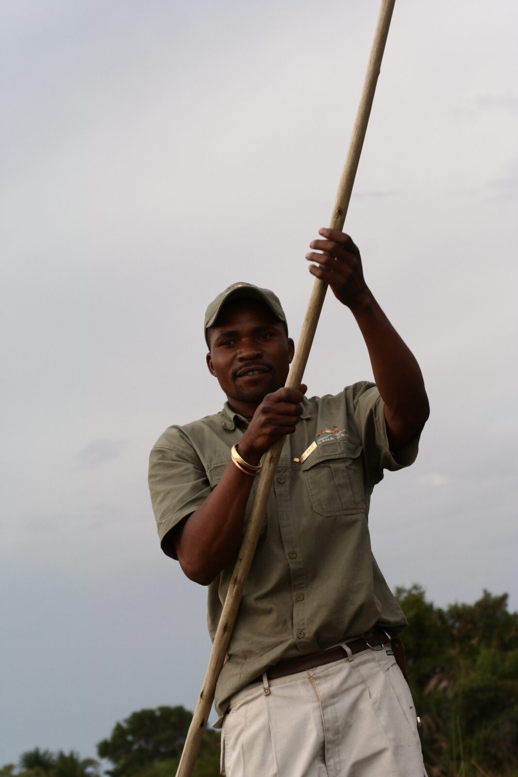 Una guida locale sorridente in divisa da safari manovra un lungo palo di legno per spingere un mokoro nei canali dell'Okavango.