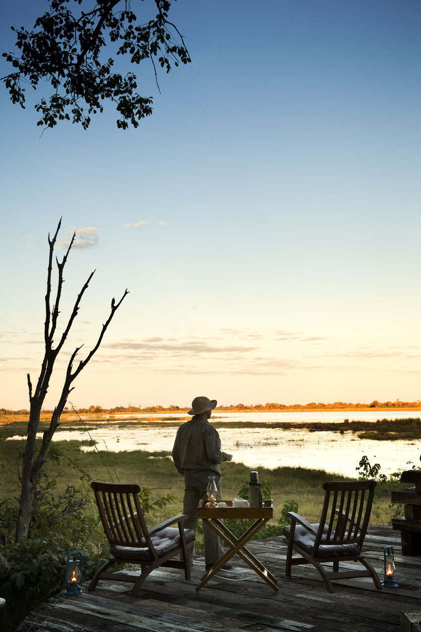 Un viaggiatore in cappello e abito da safari osserva il fiume Okavango al tramonto dalla terrazza in legno di un lodge privato in Botswana.