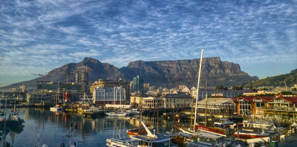 Vista del porticciolo del Victoria and Alfred Waterfront a Citta del Capo con imbarcazioni che si riflettono nell'acqua e la Table Mountain con cielo nuvoloso sullo sfondo.