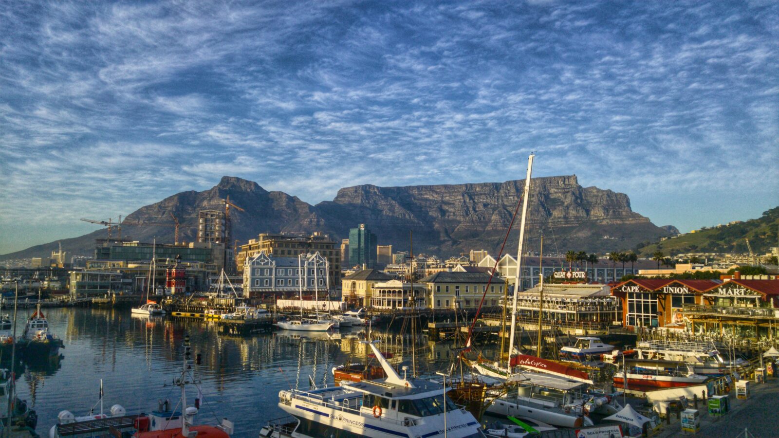 Vista del porticciolo del Victoria and Alfred Waterfront a Citta del Capo con imbarcazioni che si riflettono nell'acqua e la Table Mountain con cielo nuvoloso sullo sfondo.